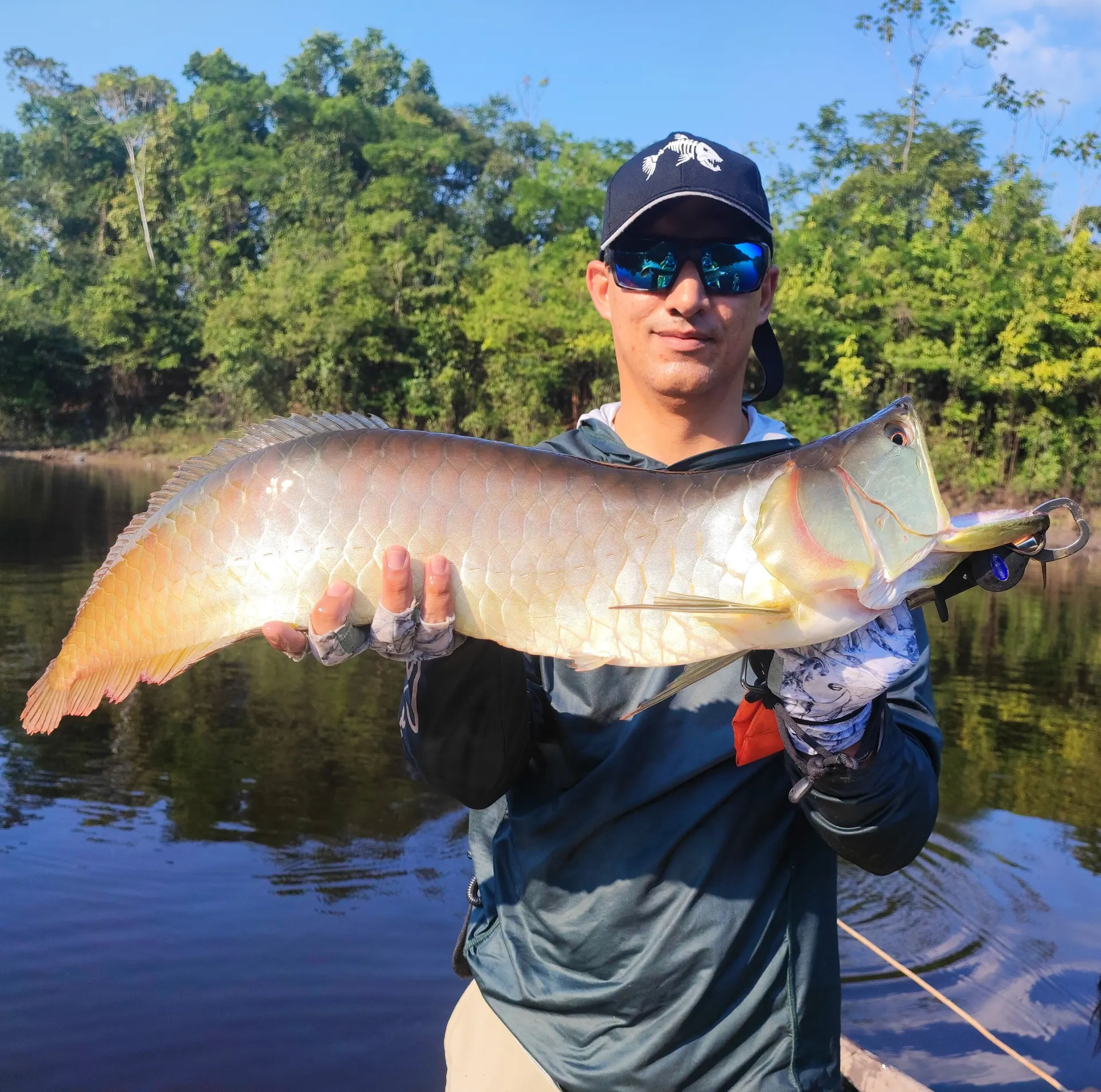 Un pescador con gorra oscura y gafas de sol sostiene una arawana grande de escamas doradas, en un río de aguas oscuras rodeado de selva.