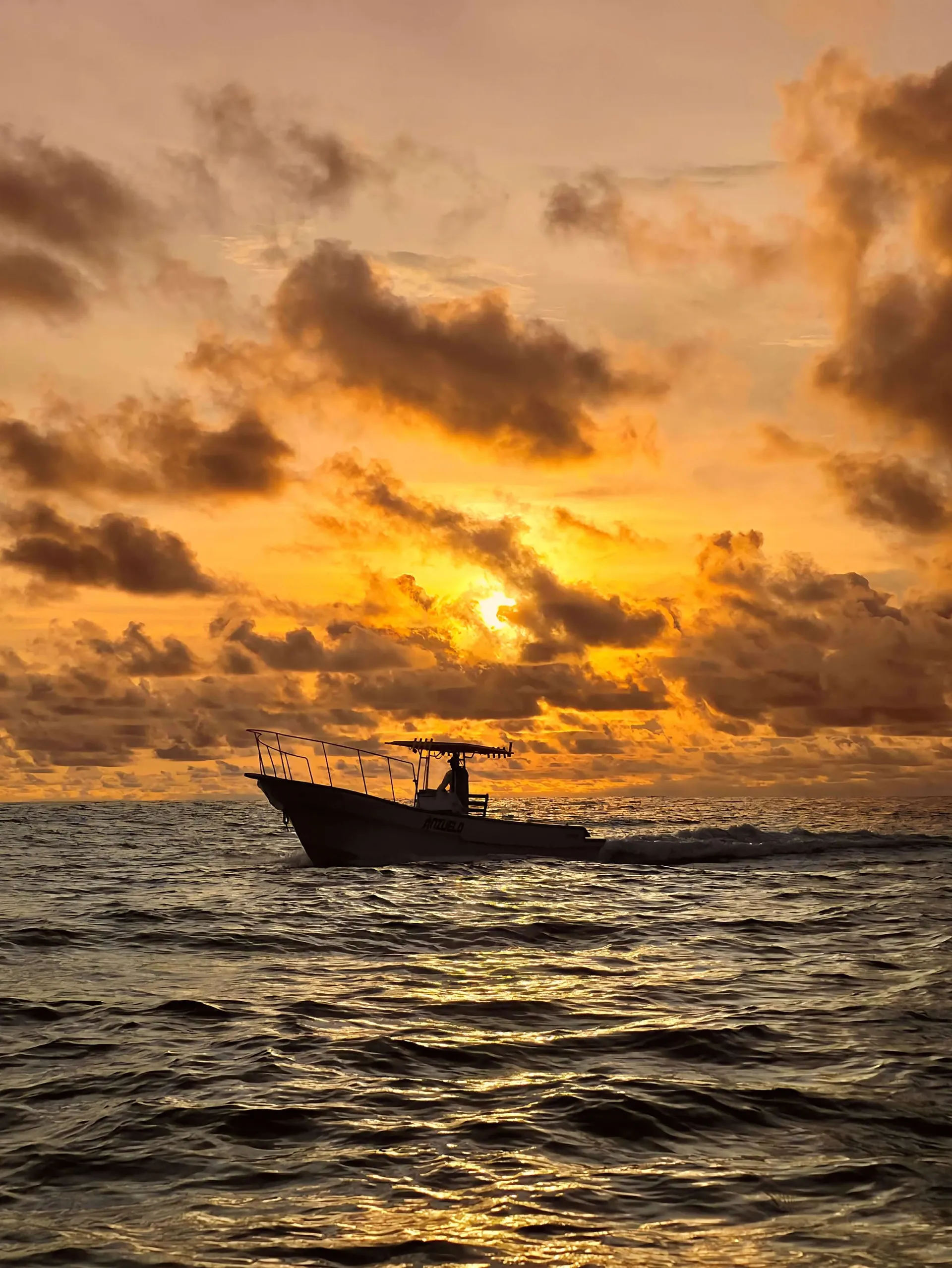 Una lancha de pesca navega en mar abierto recortada contra un amanecer anaranjado lleno de nubes.